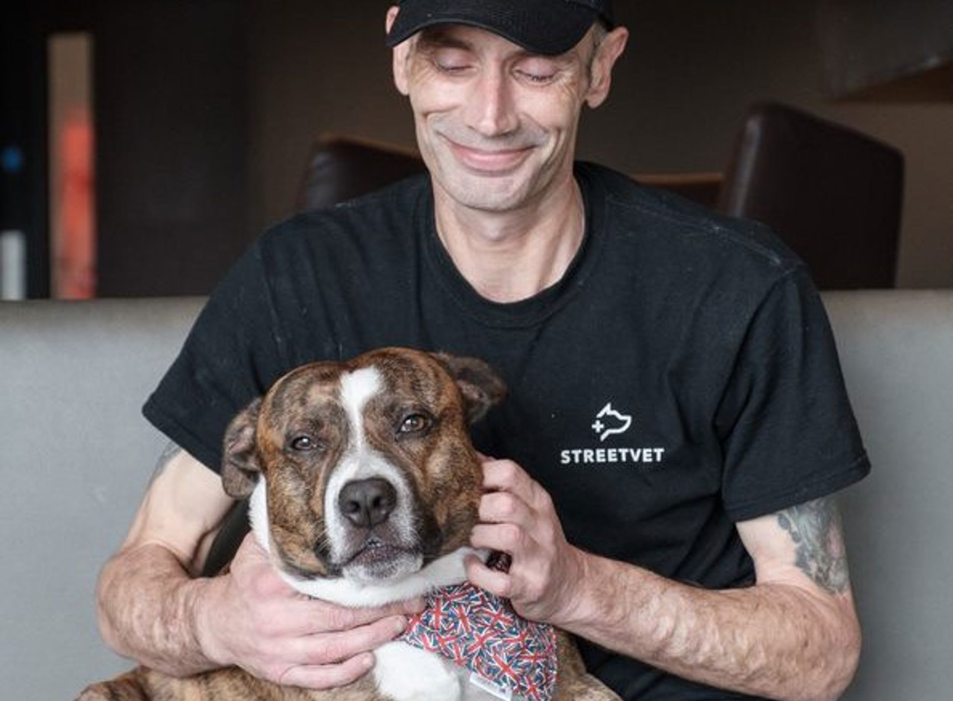 A person wearing a black StreetVet cap and black StreetVet T shirt sits indoors on a bench seat, holding a brindle dog on their lap. The dog wears a patterned bandana and faces forward, while the person’s hands gently rest around the dog’s neck and chest. The background shows a softly lit interior space with dark seating and a neutral coloured wall.
