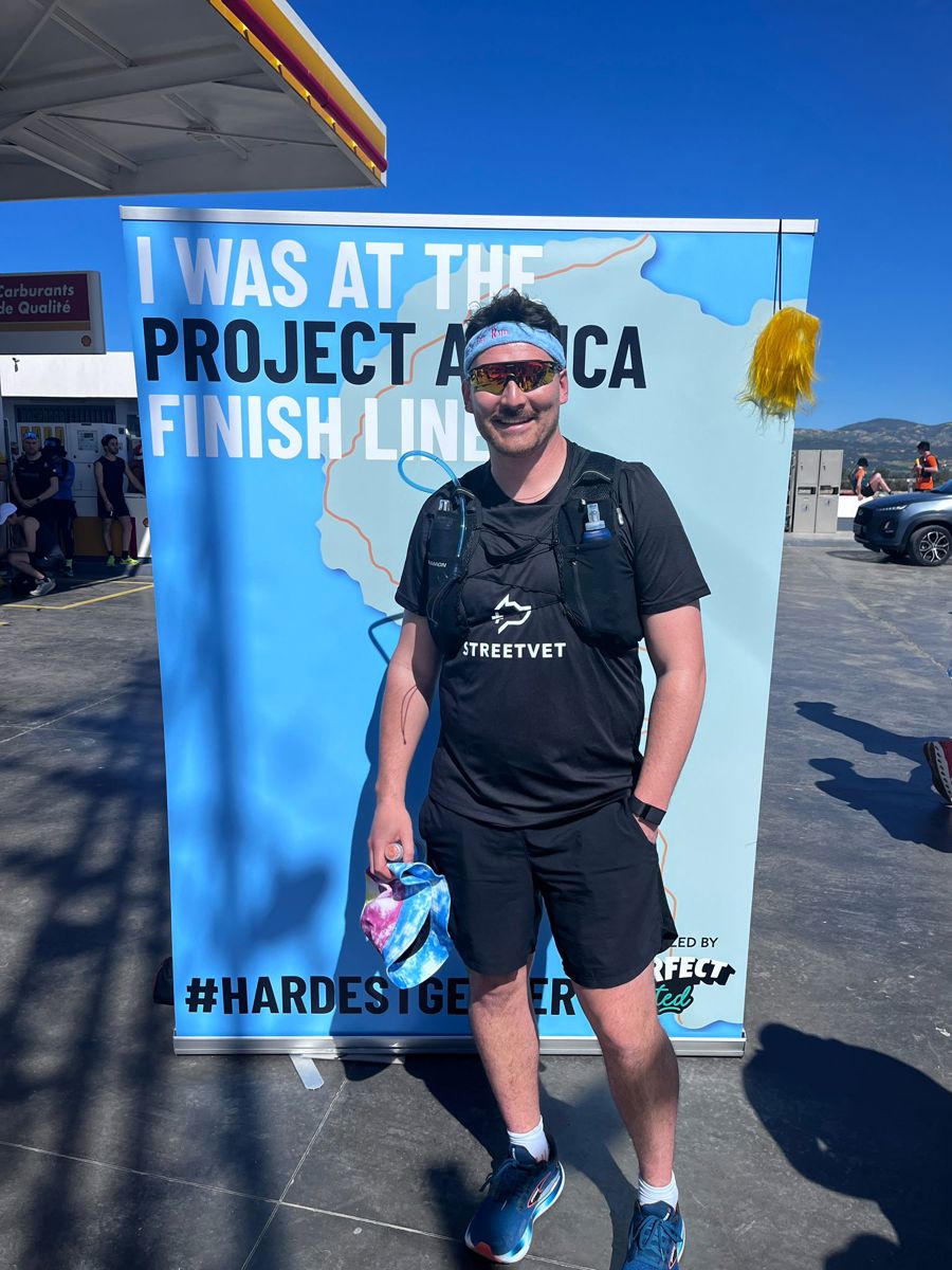 A person wearing a black StreetVet T shirt, black shorts, running shoes, sunglasses, and a hydration pack stands outdoors on a sunny day in front of a large blue and white finisher backdrop. The backdrop features an outline of a map and the text “I was at the Project Africa finish line” and “#HARDESTGEZER”. The person holds a finisher medal and a small item in one hand, with buildings, parked vehicles, and a clear blue sky visible in the background.