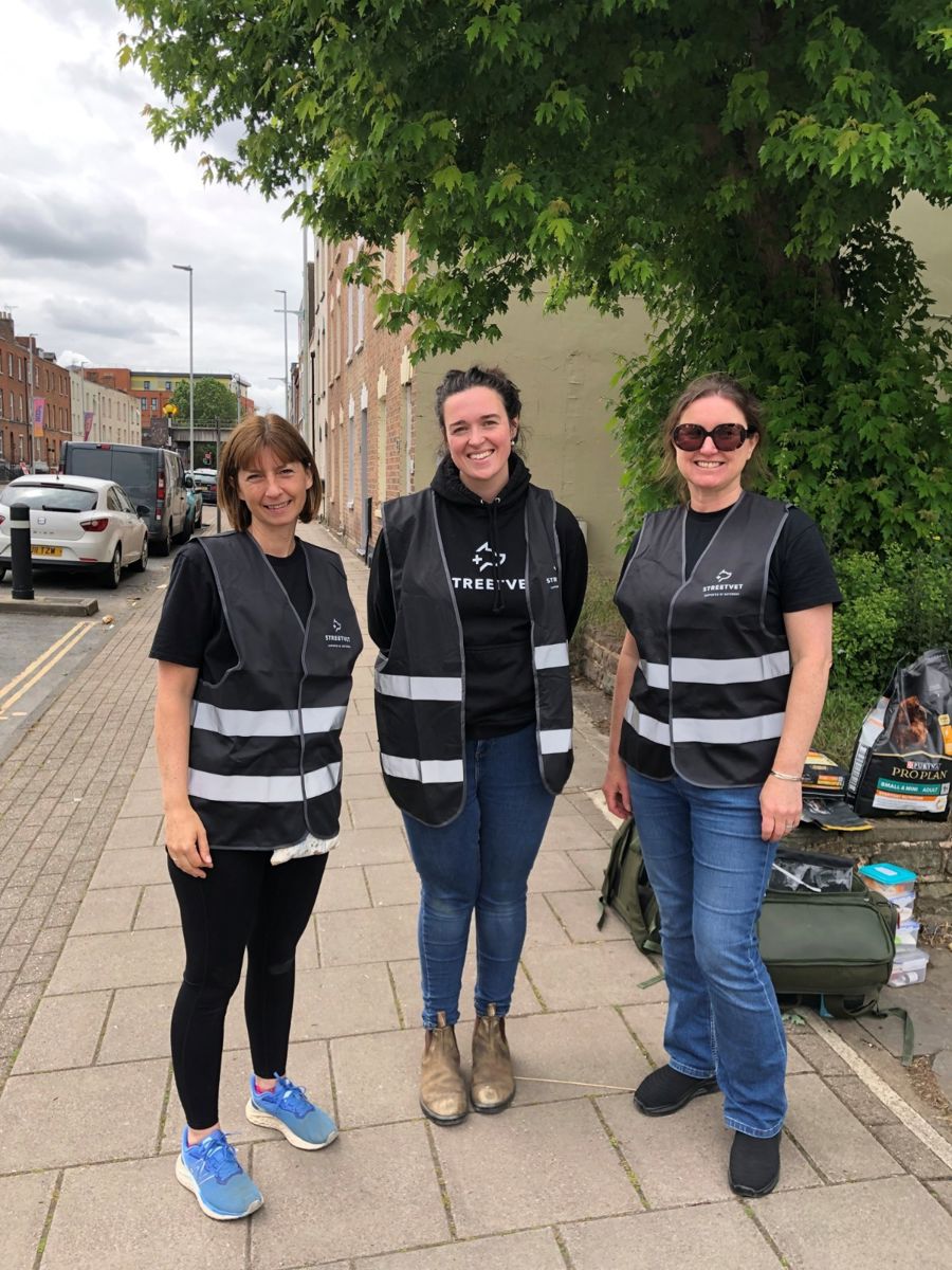 Three StreetVet volunteers stand together on a pavement in a residential street setting, wearing black clothing with reflective StreetVet vests. A tree and parked cars line the background, and veterinary equipment bags are placed on the ground nearby, indicating preparation for outreach care.