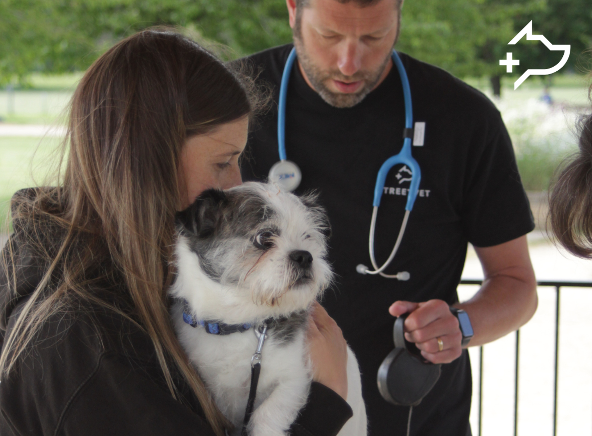 A person holds a small white and grey dog wearing a collar during an outdoor veterinary check. Another person wearing a black StreetVet T shirt with a stethoscope around the neck stands nearby, preparing or examining equipment. The setting appears to be a covered outdoor area with a railing and greenery visible in the background.
